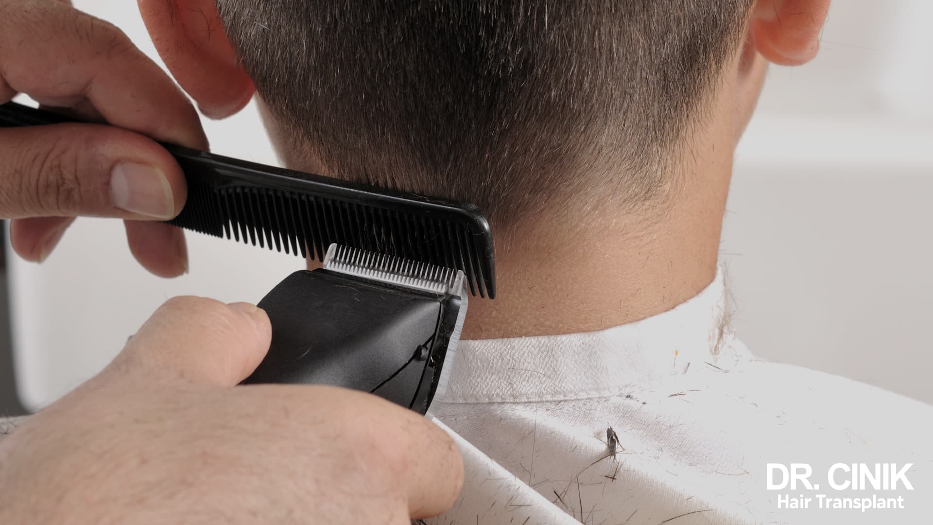 A barber running clippers along a comb at the nape, with cut hair scattered over the white cape