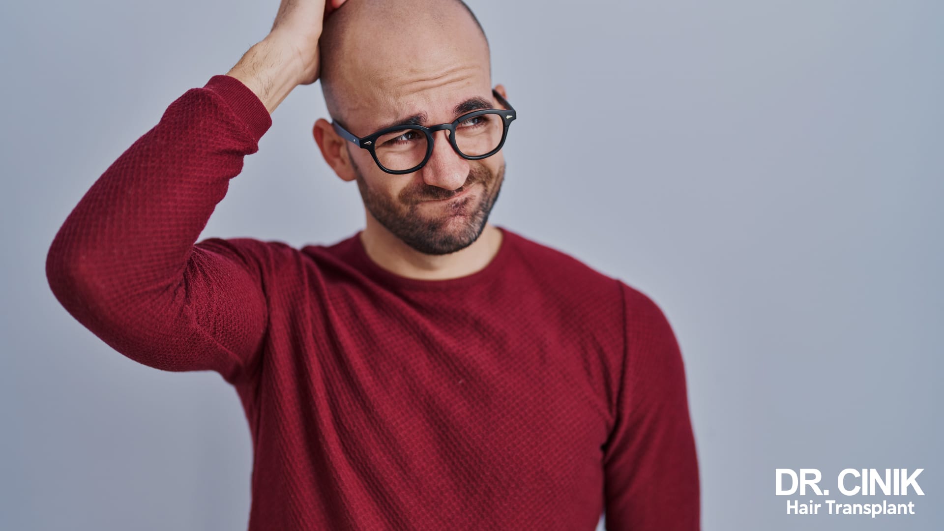 Man with advanced baldness, hand resting on the top of his head, looking puzzled