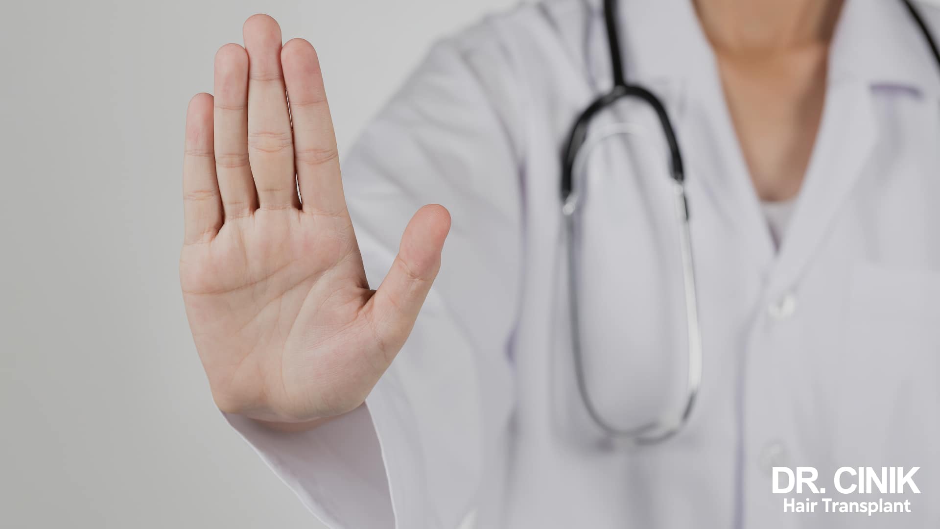 A healthcare professional in a white coat, stethoscope round the neck, raises a palm in a stop gesture