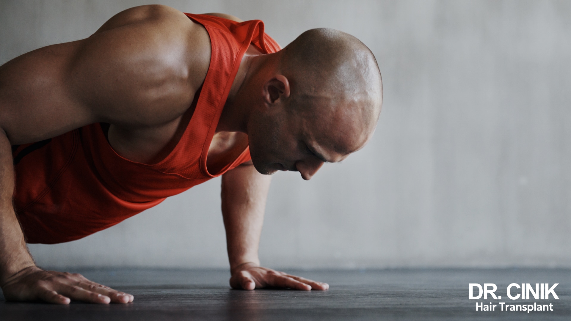 A man with a shaved head doing a press-up on the floor, in an orange vest