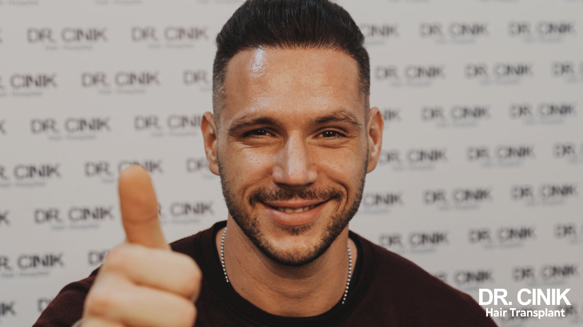 Smiling patient with thick, styled hair, giving a thumbs up in front of a wall carrying the clinic logos