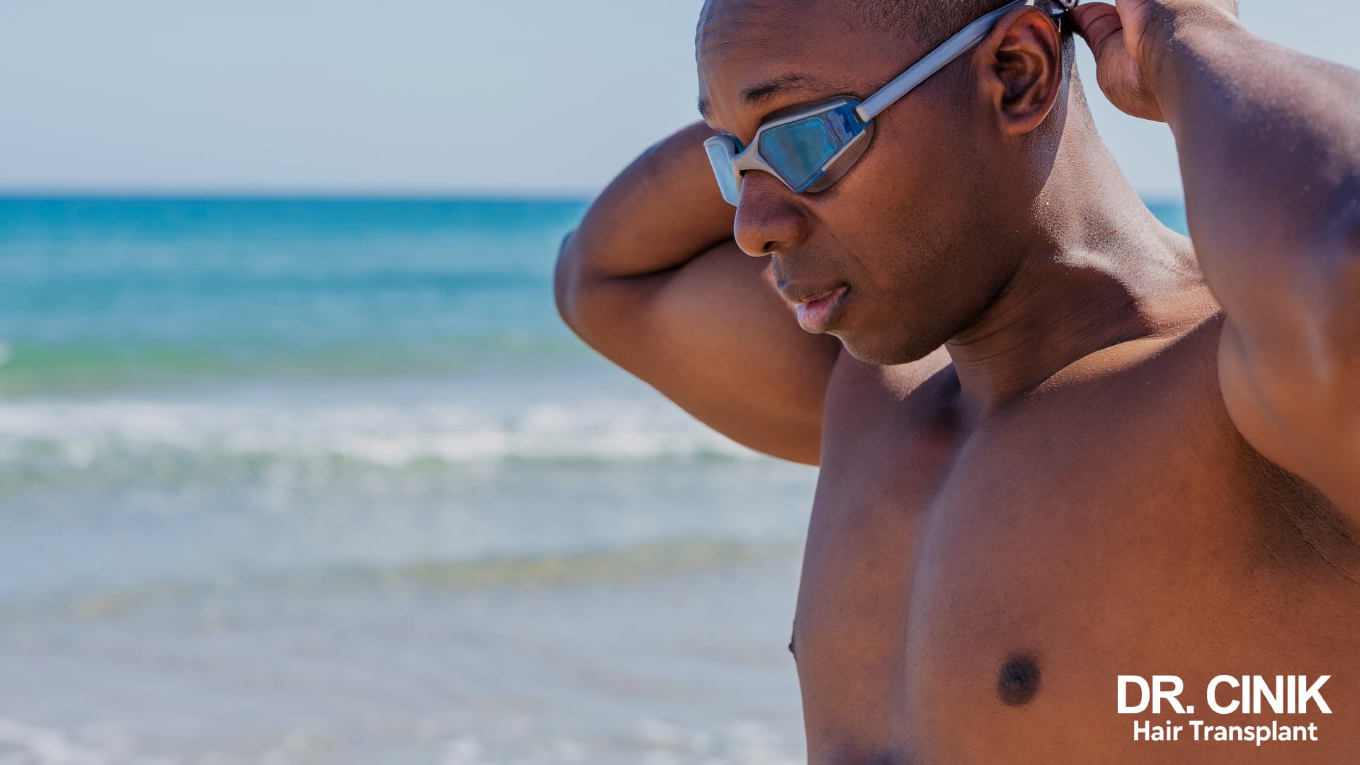 A shirtless man on a beach adjusting his swimming goggles, the sea behind him