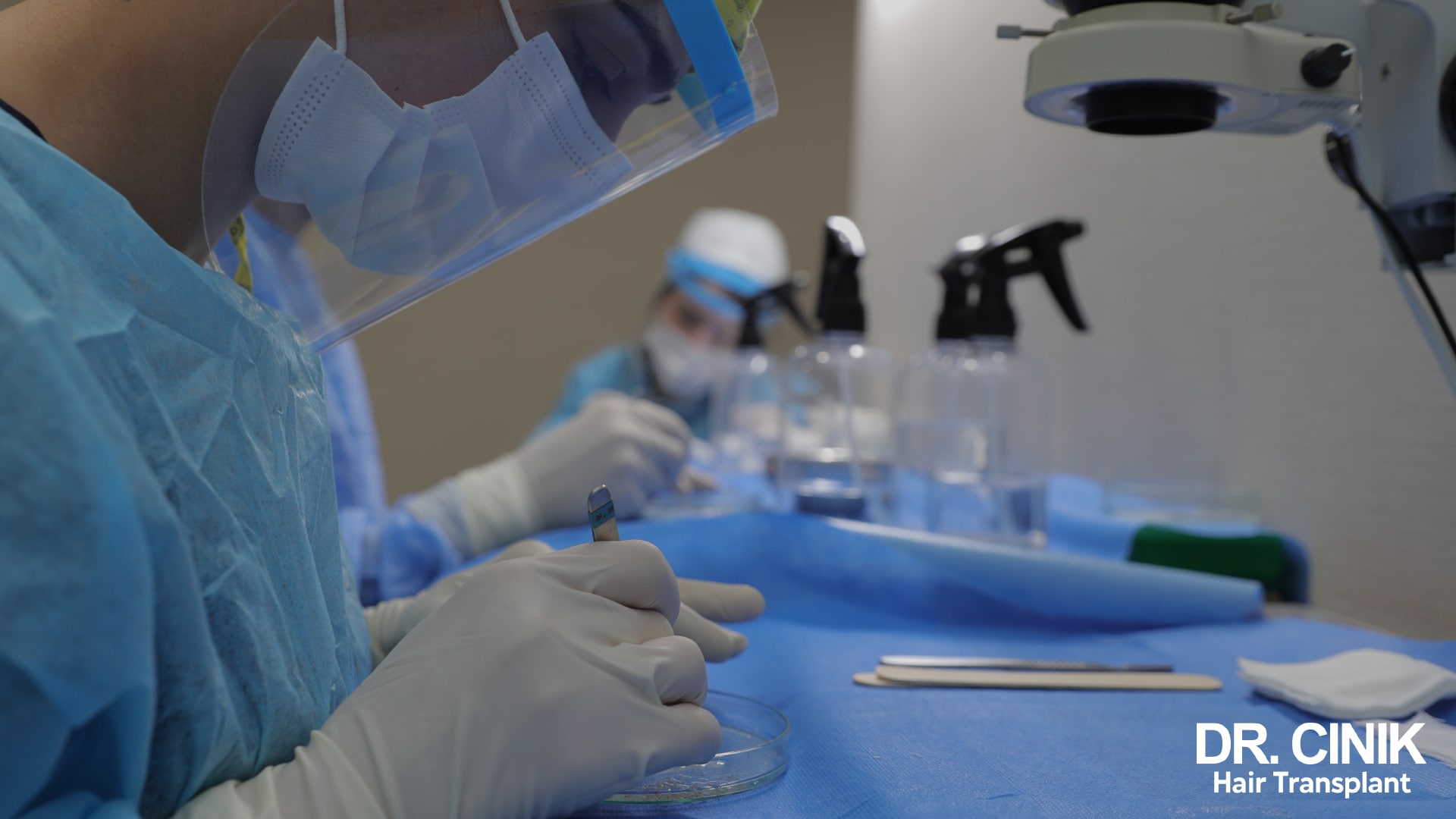 Team member in sterile clothing holding an instrument above a Petri dish, graft sorting microscope in the background