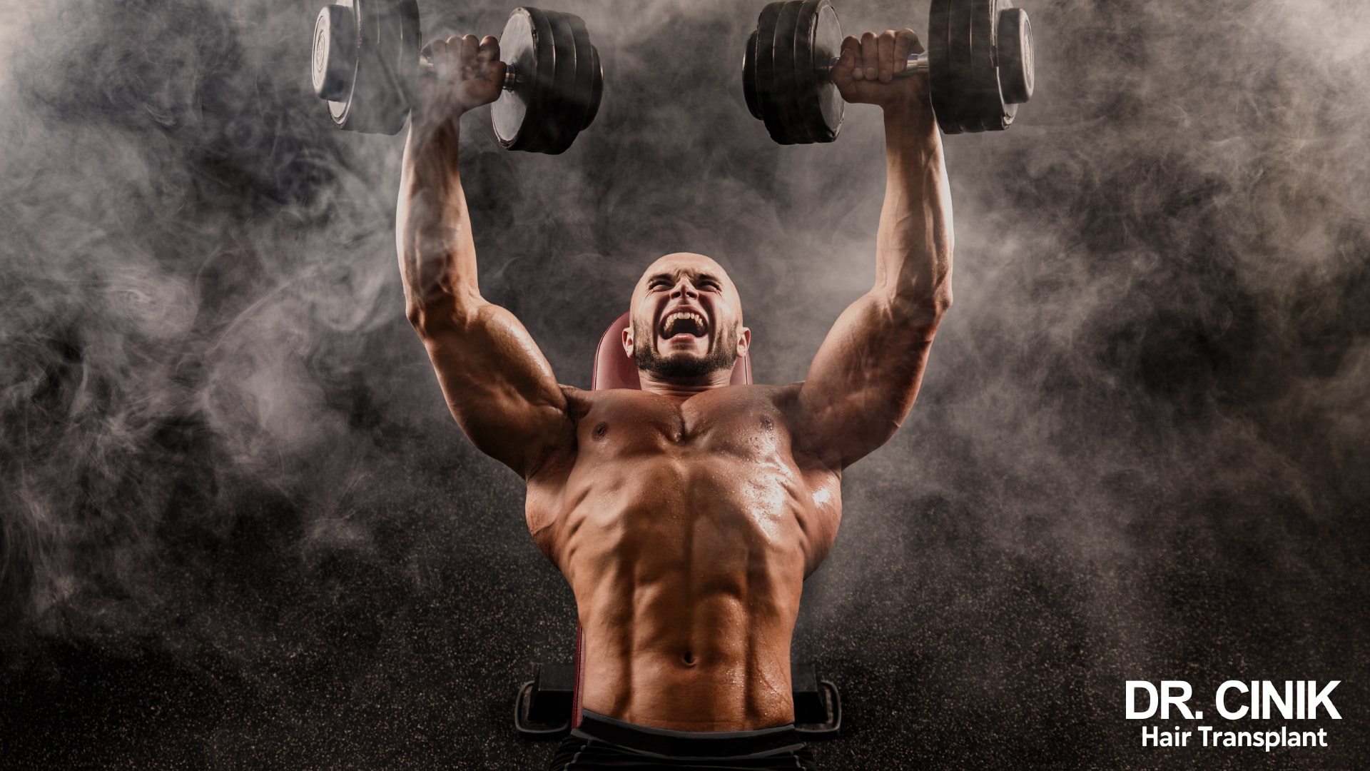 A shirtless man presses two heavy dumbbells in a dimly lit gym