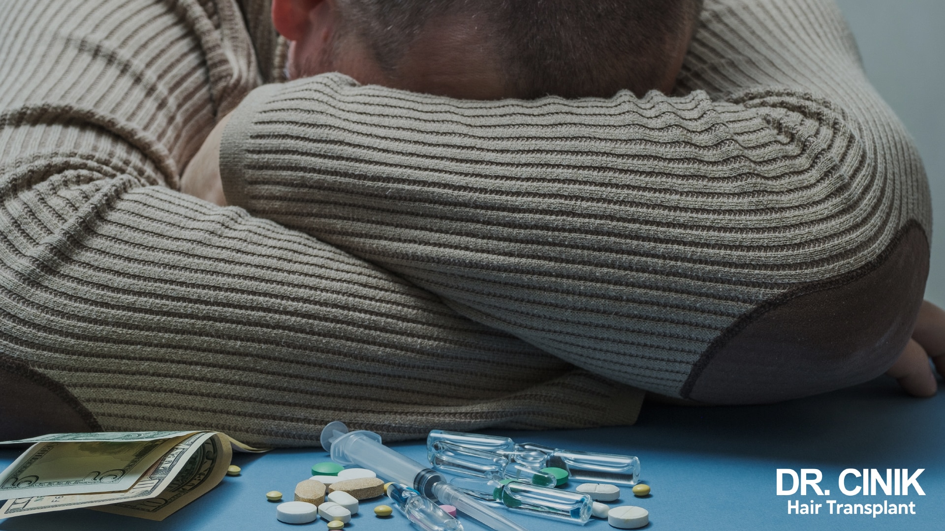 Man with his face hidden in his folded arms on a table, surrounded by tablets, vials and syringes