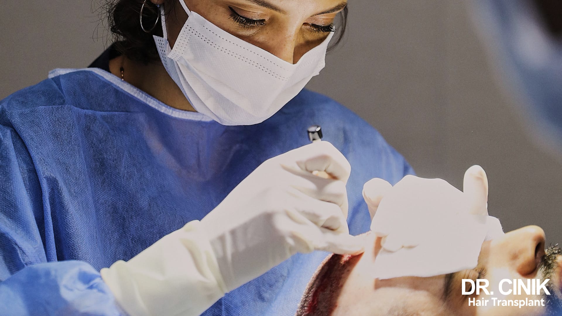 A practitioner in surgical scrubs leaning over the frontal hairline of a patient lying down, a slim instrument held between thumb and index finger