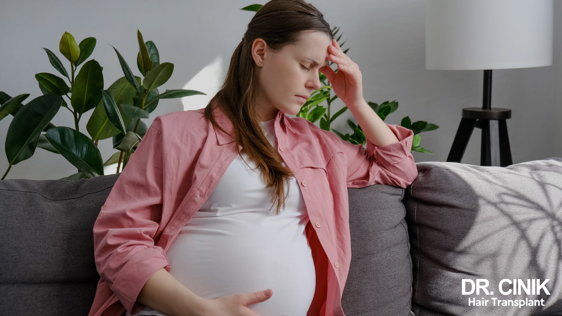Pregnant woman sitting on a sofa, one hand on her forehead and the other on her belly, eyes closed