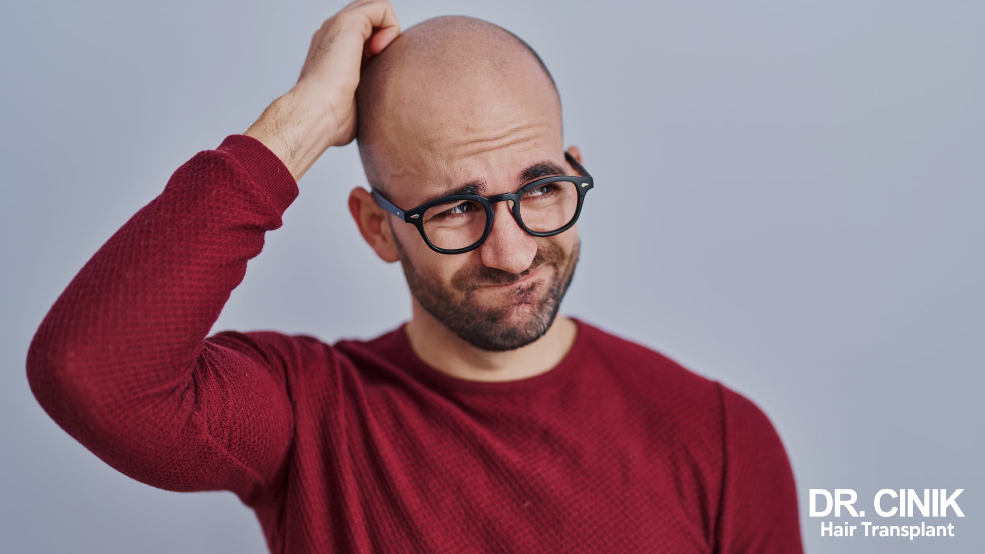 Bald man in glasses scratching the top of his head with a puzzled expression
