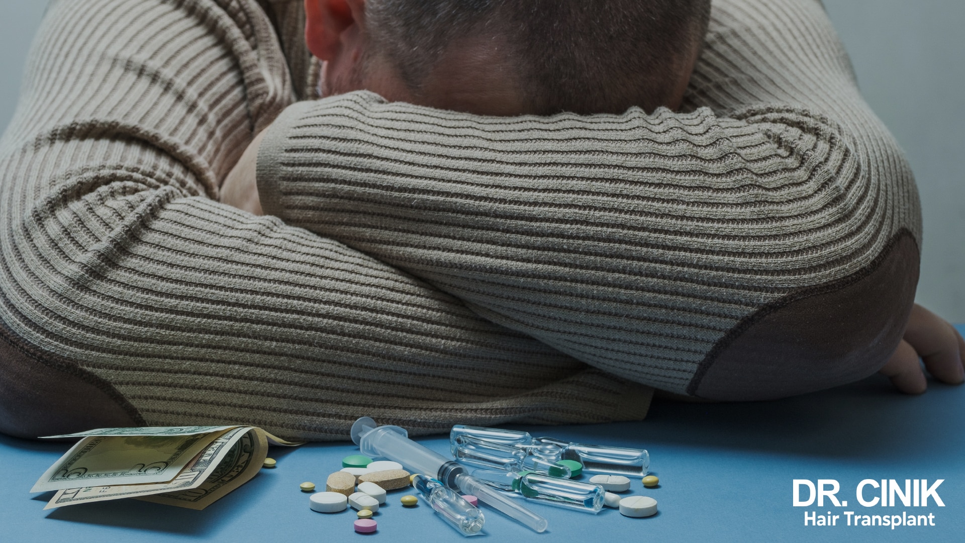 Man slumped over a table, head on his arms, surrounded by tablets, blister packs and vials