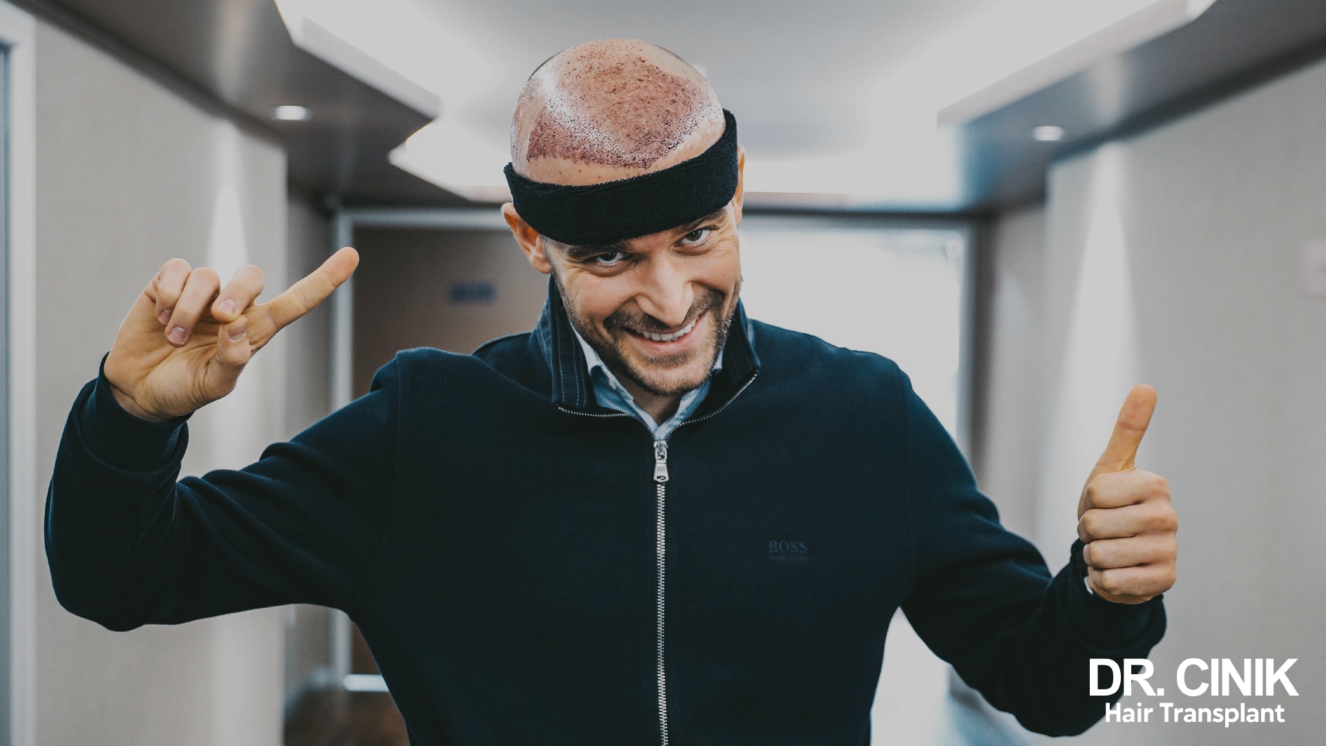 Patient just out of surgery, black headband on the forehead, giving a victory sign and a thumbs up in a corridor, recipient area red and covered with fresh grafts
