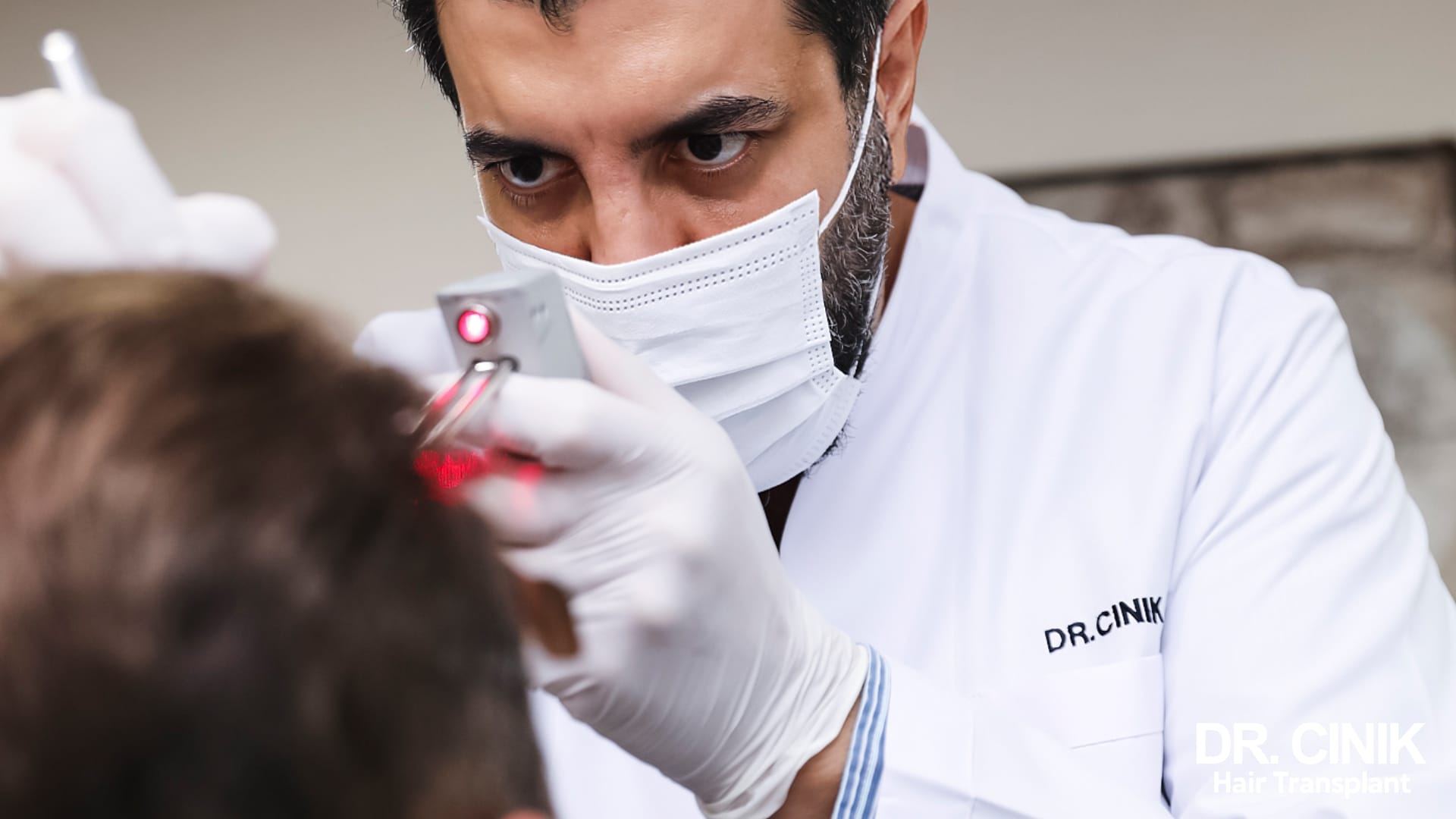 Dr Cinik, in scrubs and mask, examining a patient's scalp with a red light device