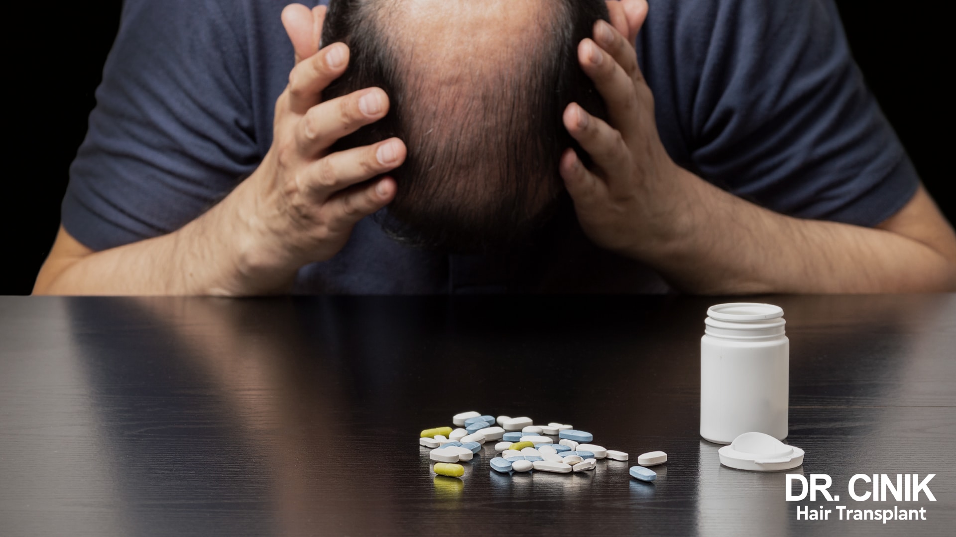 Balding man with his head in his hands in front of tablets scattered on a table
