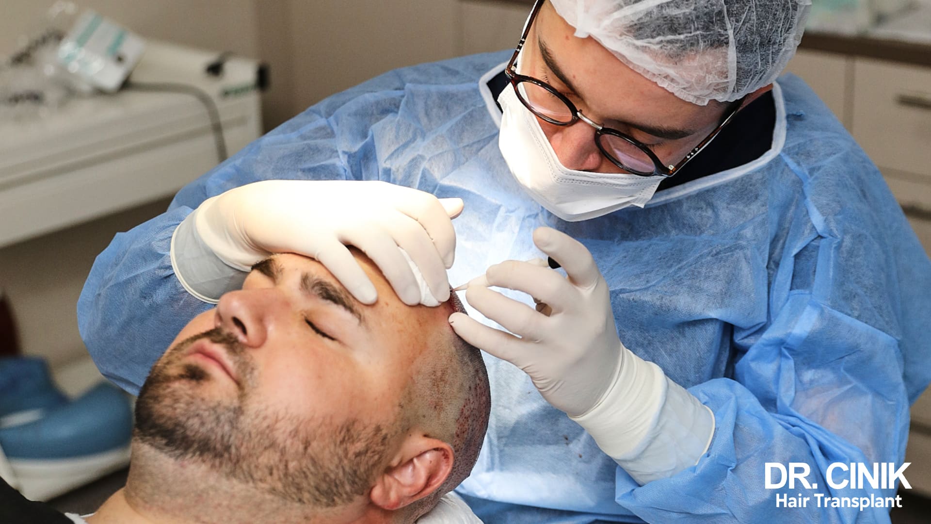 Practitioner in sterile scrubs, cap and gloves implanting grafts along the hairline of a patient lying in the operating room