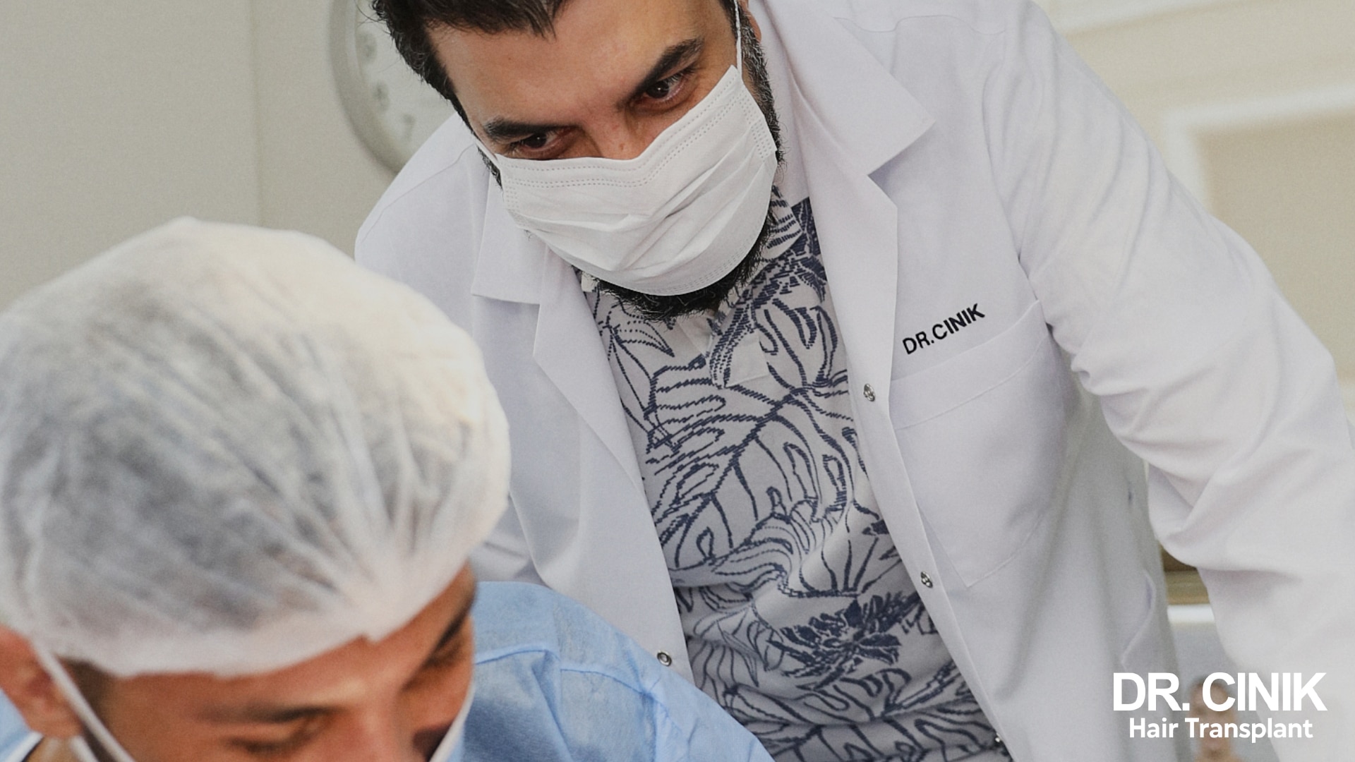 Dr Emrah Cinik in a white coat and mask, leaning over a patient wearing a surgical cap in the operating room