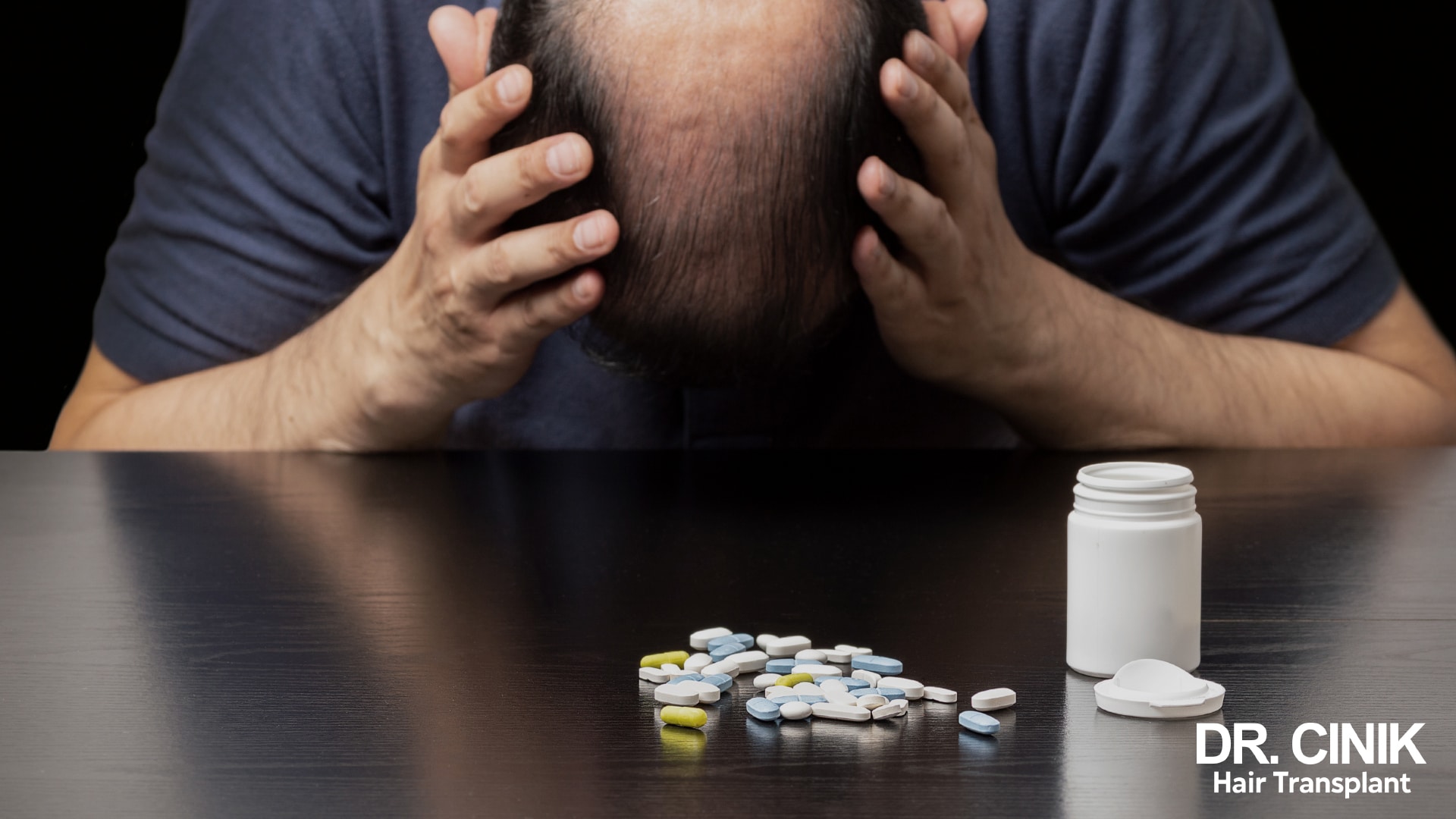 Man balding at the crown sitting at a dark table, head in his hands, in front of an open bottle and scattered tablets