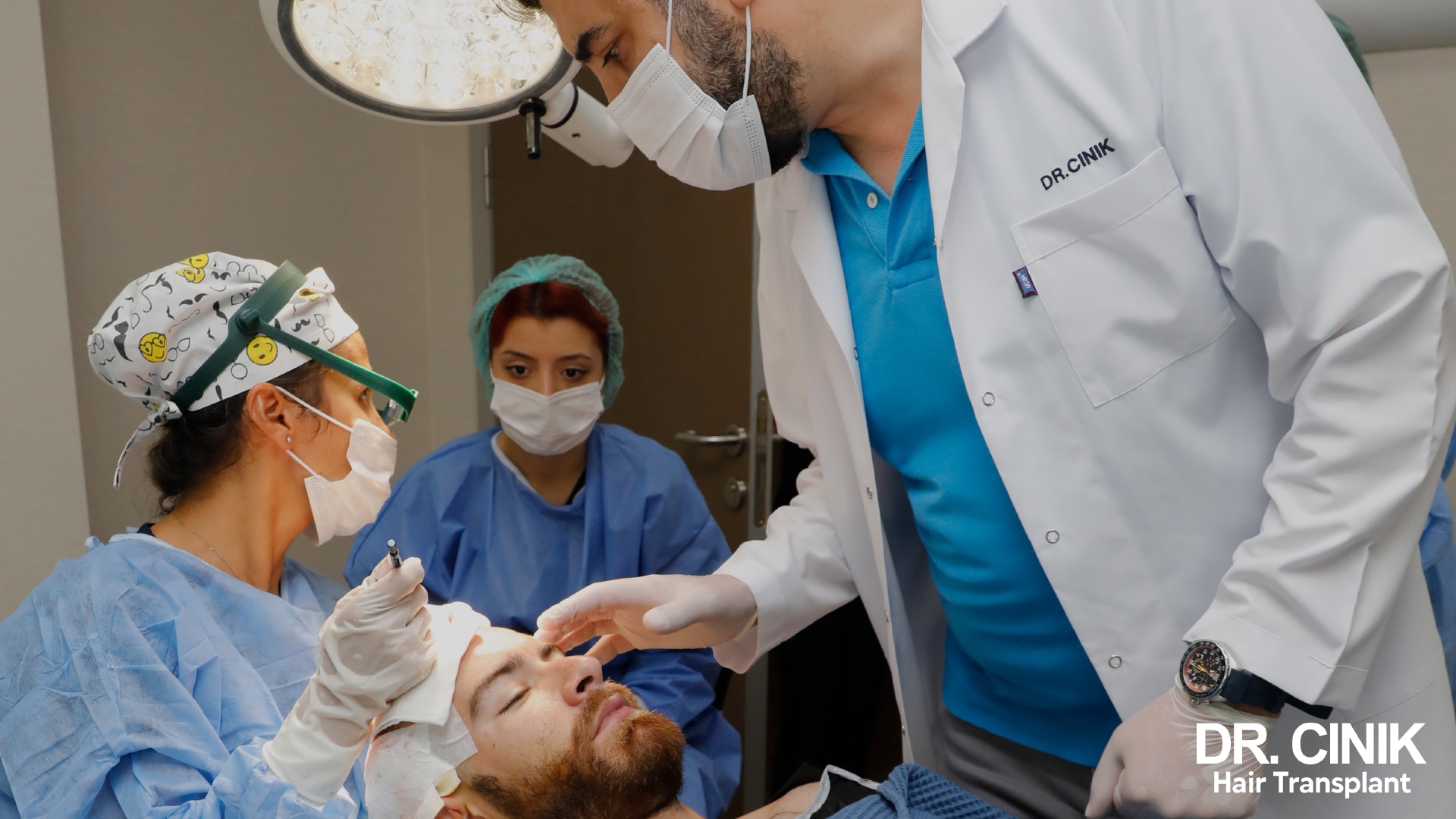 Chirurgien en blouse blanche portant une loupe frontale, penché au-dessus d'un patient allongé et assisté de deux collègues en tenue chirurgicale