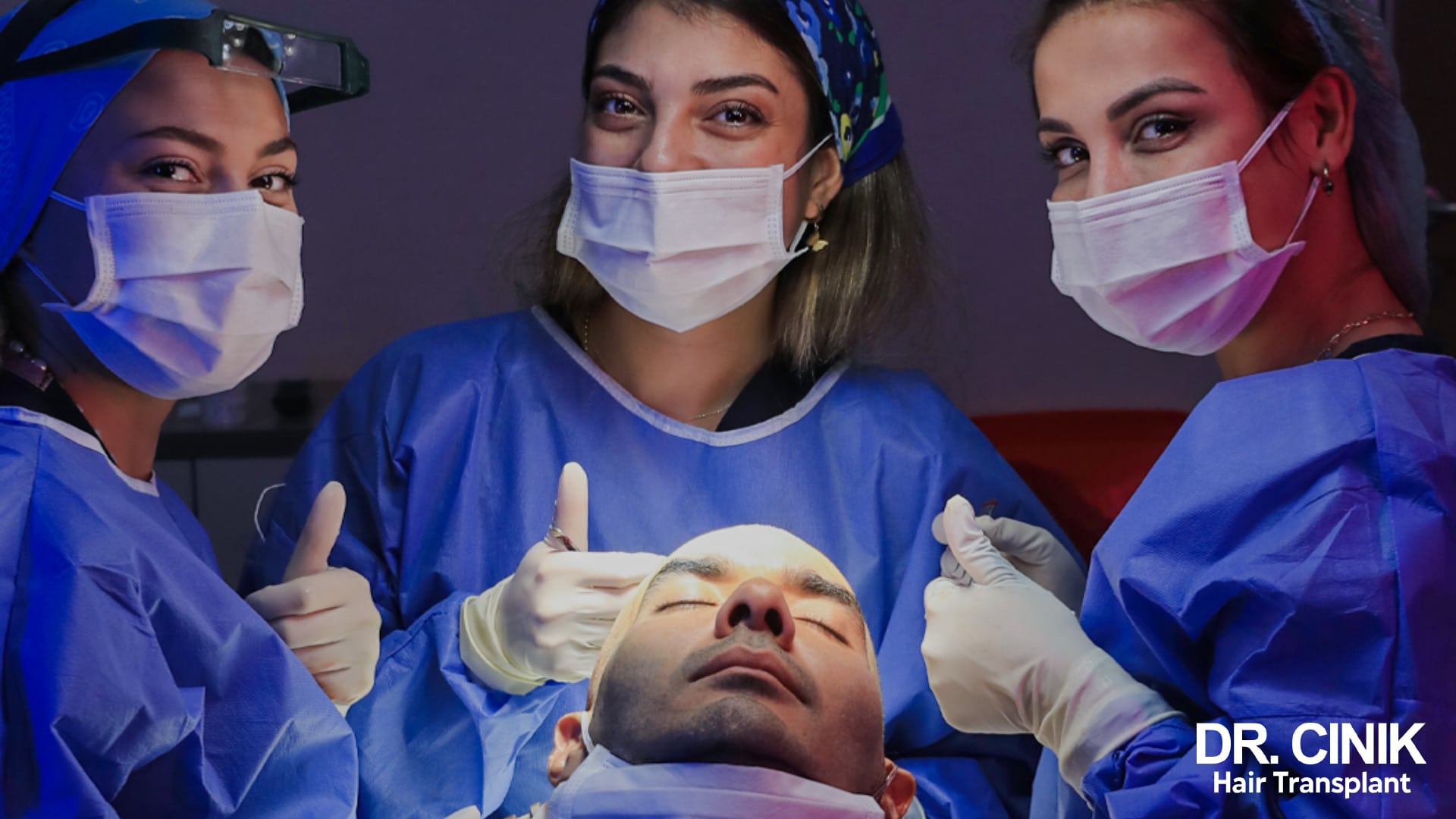Trois soignantes en tenue chirurgicale et masques autour d'un patient allongé sous une lampe opératoire, deux d'entre elles faisant un signe pouce levé