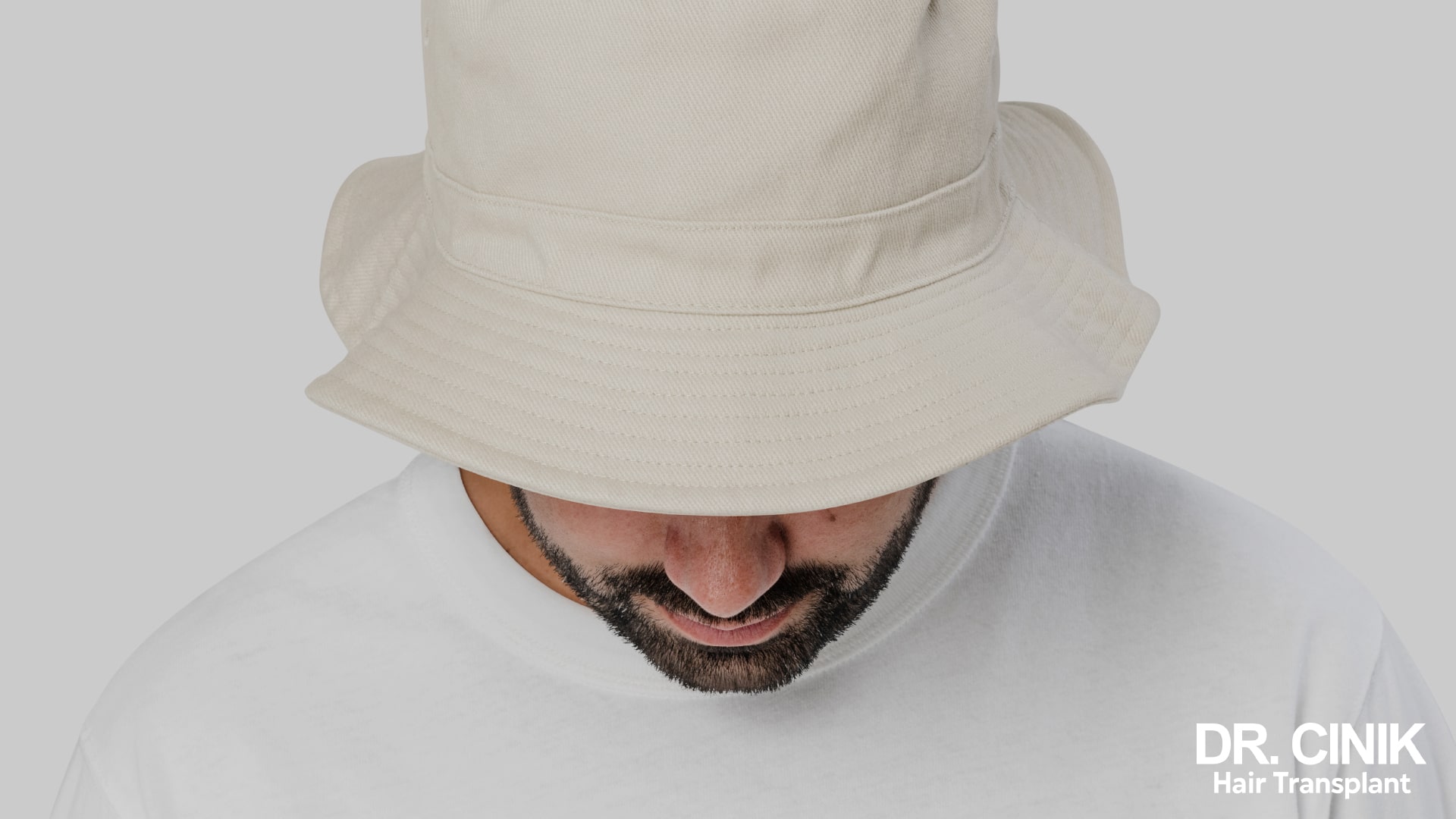 A man seen from above, head bowed, wearing a soft ecru bucket hat whose wide brim covers the entire skull