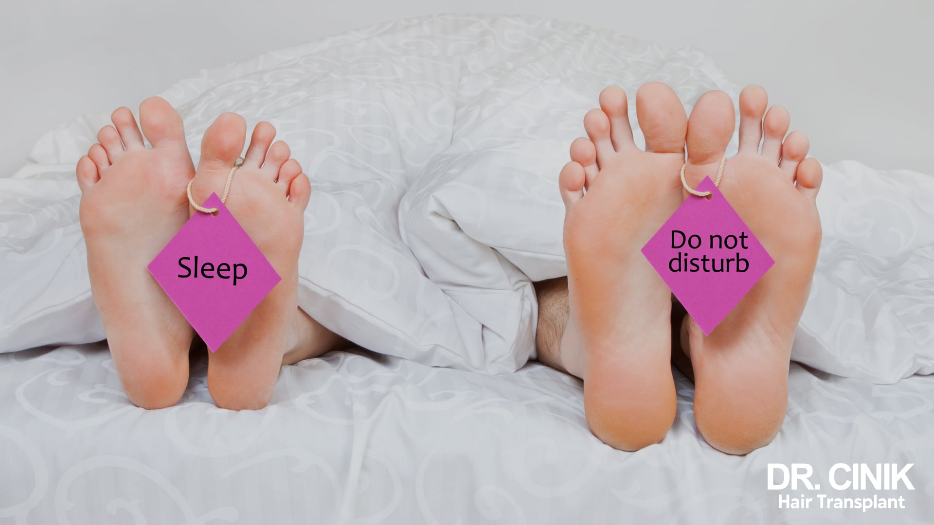 Two people lying under a duvet with only their feet showing, each wearing a humorous tag reading Sleep and Do not disturb