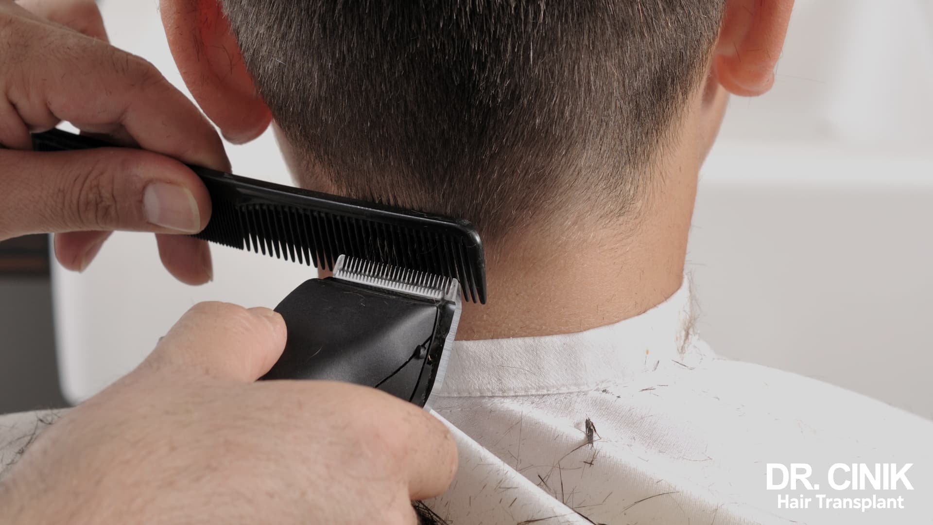 Barber shaving the nape of a man's neck with clippers and a comb, cut hair falling onto a white cape